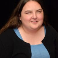 Medium shot headshot of Claire Stanley on a black background. Claire is wearing a blue dress shirt with a black sweater.